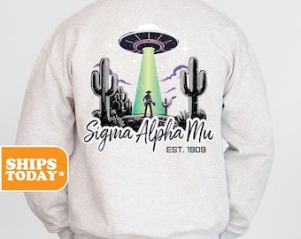May include: A grey sweatshirt featuring a graphic of a UFO hovering over a desert scene with cacti and a cowboy. The text "Sigma Alpha Mu Est. 1909" is printed beneath the graphic.