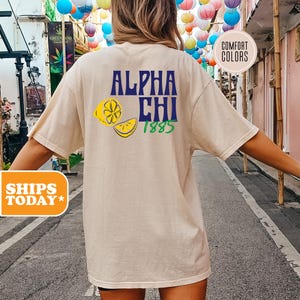 May include: A beige short-sleeve t-shirt featuring a graphic on the back with the text ALPHA CHI in blue, the number 1885 in green, and two yellow lemon slices. A circular badge on the upper right corner contains the text COMFORT COLORS.
