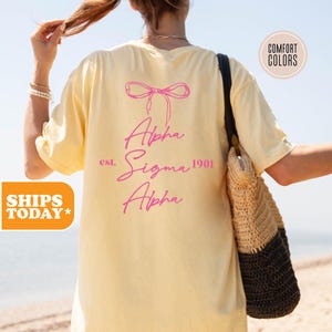 May include: A yellow t-shirt with a pink bow and the text "Alpha Sigma Alpha est. 1901" on the back.