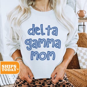 May include: White sweatshirt with the blue text "Delta Gamma Mom". The text has a textured, sketched appearance. The model is wearing leopard print leggings and a gold chain necklace.