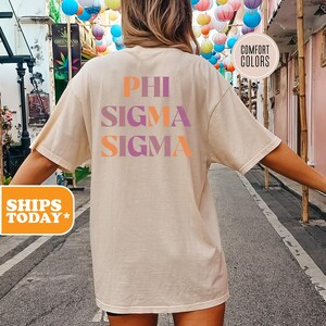 May include: A beige t-shirt with the words "PHI SIGMA SIGMA" printed on the back in orange and purple. The shirt is made of a soft material and is a Comfort Colors brand.