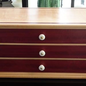 May include: A wooden jewelry box with three drawers. The box features a light-colored top and base, with a dark red-brown body and drawers. Each drawer has a white knob with a gold accent. The box is approximately 12 inches wide.