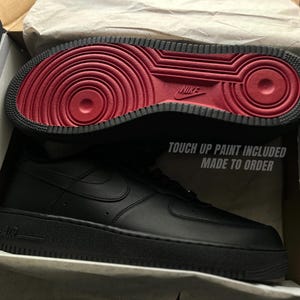 May include: Black leather sneaker with a red sole, displayed in an open box. The sole features a circular pattern and the Nike logo. The text "TOUCH UP PAINT INCLUDED MADE TO ORDER" is visible on the box.