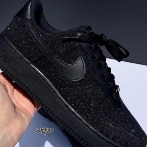 May include: Black glitter sneaker with black laces and a black Nike swoosh. The shoe has a black sole and the word "AIR" is visible on the side. The text "custom snkrs" is also visible on the side of the shoe.