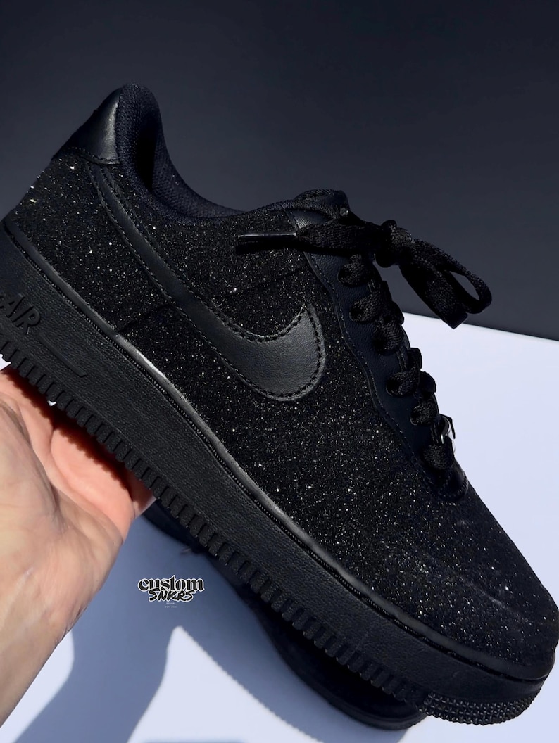 May include: Black glitter sneaker with black laces and a black Nike swoosh. The shoe has a black sole and the word "AIR" is visible on the side. The text "custom snkrs" is also visible on the side of the shoe.