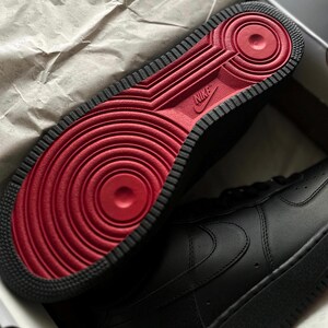 May include: Black Nike Air Force 1 sneakers with a red sole. The sole features a circular design and the Nike logo. The sneakers are in a box with the text "custom snkrs".