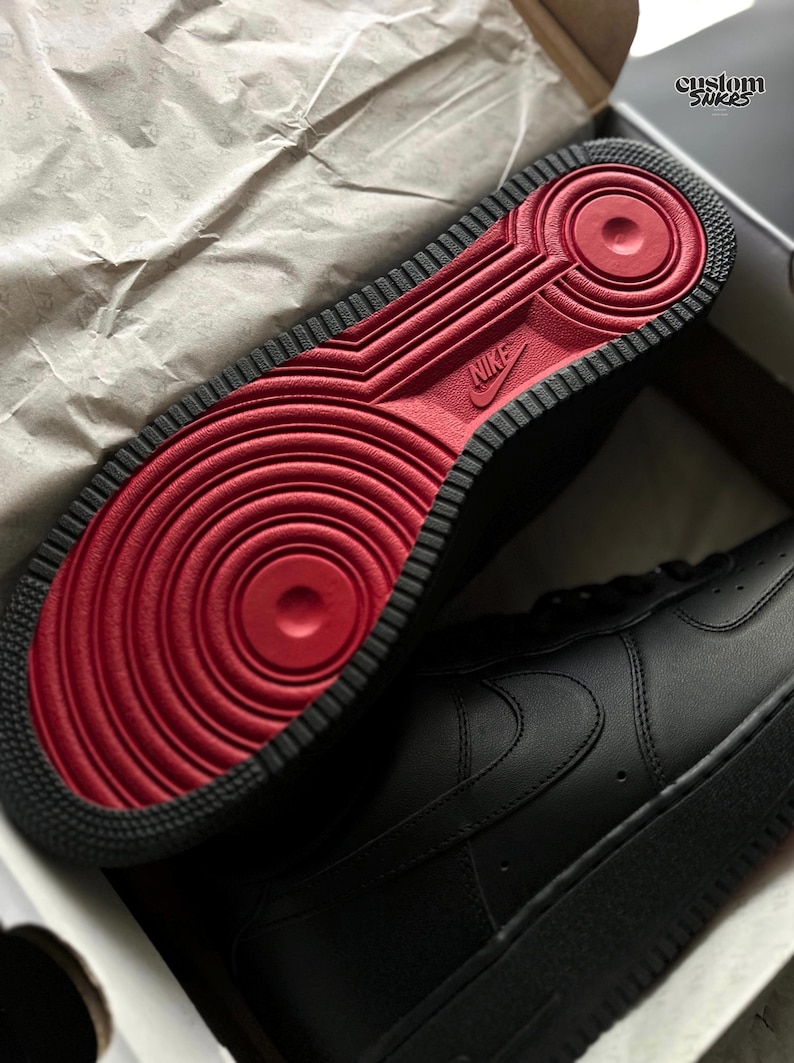 May include: Black Nike Air Force 1 sneakers with a red sole. The sole features a circular design and the Nike logo. The sneakers are in a box with the text "custom snkrs".
