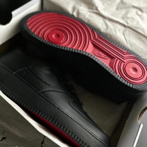 May include: Black Air Force 1 sneakers with red soles, presented in a box. The sneakers have a smooth leather upper, a classic design, and the Nike swoosh logo on the side. The red soles provide a striking contrast to the black.