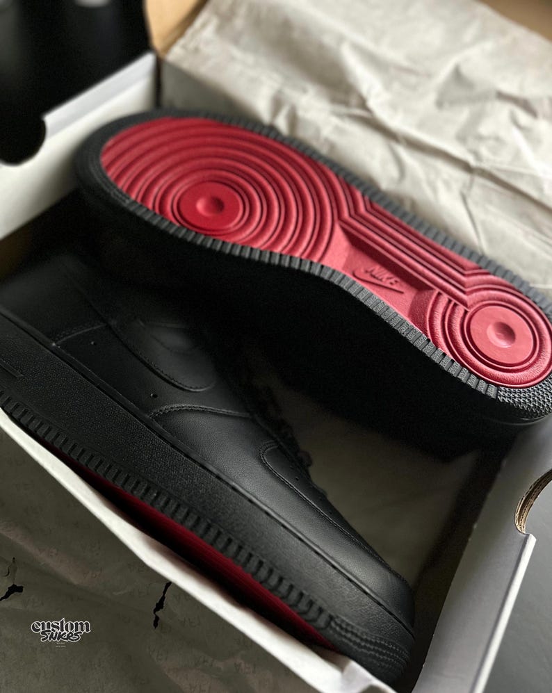 May include: Black Air Force 1 sneakers with red soles, presented in a box. The sneakers have a smooth leather upper, a classic design, and the Nike swoosh logo on the side. The red soles provide a striking contrast to the black.