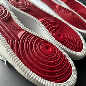 May include: Close-up of the soles of white sneakers with red rubber bottoms. The soles have a circular pattern and the Nike logo is visible. The text "custom snkrs" is in the lower left corner. The sneakers are on a dark surface.