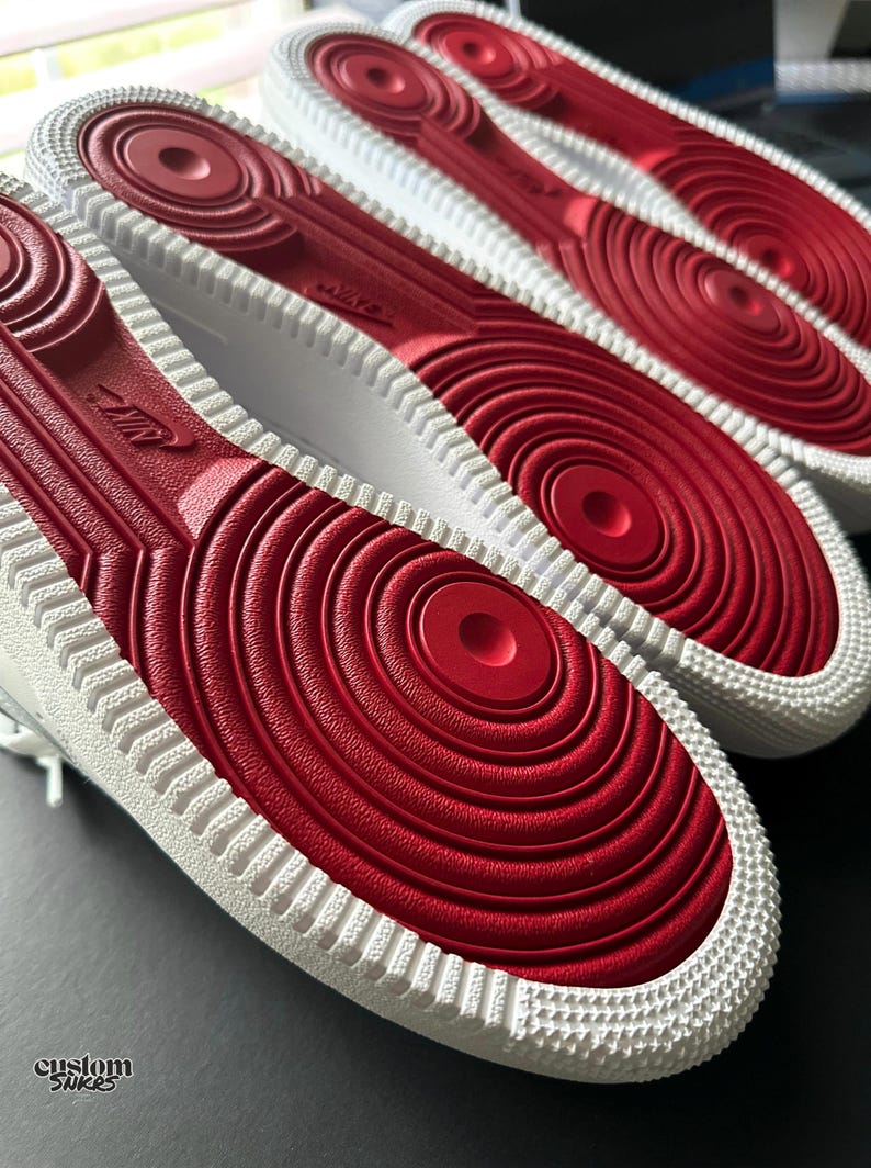 May include: Close-up of the soles of white sneakers with red rubber bottoms. The soles have a circular pattern and the Nike logo is visible. The text "custom snkrs" is in the lower left corner. The sneakers are on a dark surface.