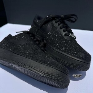 May include: Pair of black glitter-covered trainers with black laces and a black Nike swoosh. The shoes are on a white surface with a dark background. The soles are black and textured. The words "custom snkrs" are visible.