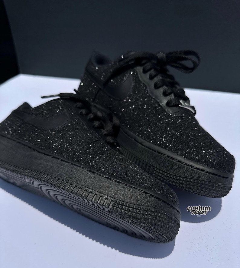 May include: Pair of black glitter-covered trainers with black laces and a black Nike swoosh. The shoes are on a white surface with a dark background. The soles are black and textured. The words "custom snkrs" are visible.