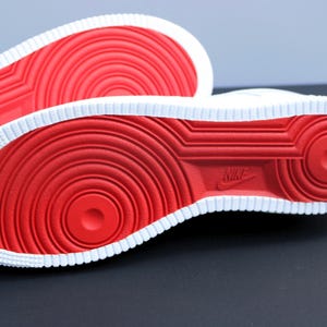 May include: Close-up of the soles of two white sneakers with red circular patterns and a white border. The Nike logo is visible on the sole. The sneakers are on a dark surface.