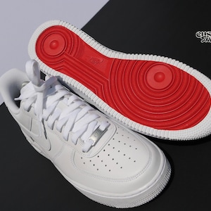 May include: White leather sneakers with a red sole. The sneakers have white laces and a silver metal lace tag. The sole is red with a circular design. The word "Nike" is visible on the sole. The text "custom sticks" is in the upper right corner.