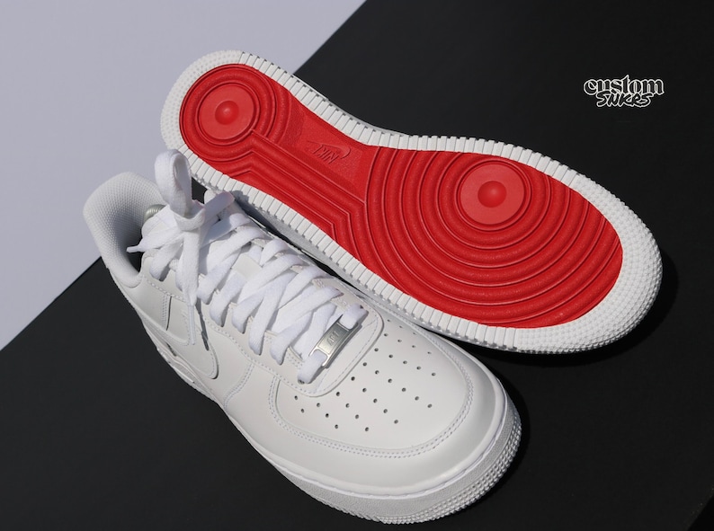 May include: White leather sneakers with a red sole. The sneakers have white laces and a silver metal lace tag. The sole is red with a circular design. The word "Nike" is visible on the sole. The text "custom sticks" is in the upper right corner.
