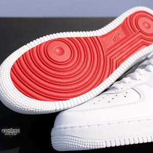 May include: White sneakers with a red sole. The sole features a circular pattern and the Nike logo. The shoe's upper is smooth white leather with a classic design. The image shows the sole and part of the shoe.