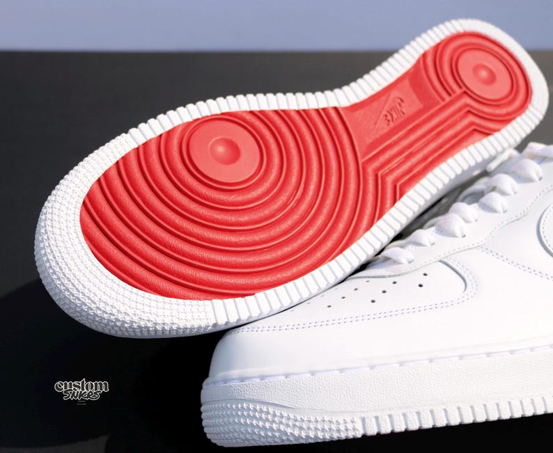 May include: White sneakers with a red sole. The sole features a circular pattern and the Nike logo. The shoe's upper is smooth white leather with a classic design. The image shows the sole and part of the shoe.