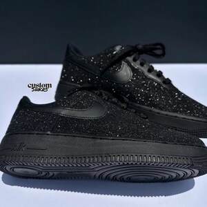May include: Black glitter-covered trainers with a classic design. The shoes feature the Nike swoosh logo and black laces. The soles are solid black. The text "custom snkrs" is visible. These shoes are a stylish footwear option.