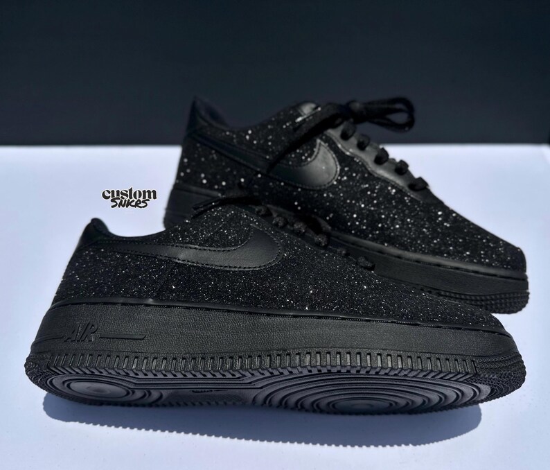 May include: Black glitter-covered trainers with a classic design. The shoes feature the Nike swoosh logo and black laces. The soles are solid black. The text "custom snkrs" is visible. These shoes are a stylish footwear option.