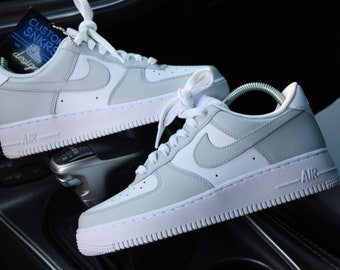 May include: A pair of white and light gray Nike Air Force 1 sneakers with white laces. The sneakers are sitting on the center console of a car. The tag on the left sneaker says "Custom SNKRs" and "design by".