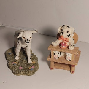 May include: Two ceramic Dalmatian dog figurines. One is standing on a green base with flowers. The other is sitting in a wooden chair with a pink heart.