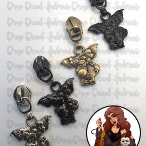 May include: Four zipper pulls featuring a cartoon monster with large ears and a toothy grin. The zipper pulls are in four different finishes: silver, black, gold, and antique silver. The zipper pulls are displayed on a white background with a cartoon illustration of a woman sewing.