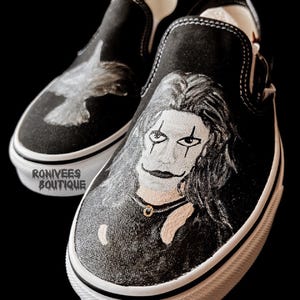 Puede incluir: Un par de zapatillas negras sin cordones con ilustraciones pintadas a mano. Un zapato muestra el retrato de una persona con maquillaje facial blanco y marcas oscuras en los ojos, el otro un ave en vuelo. El texto RONIVEES BOUTIQUE aparece en el lateral.