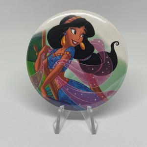 May include: A round button featuring a cartoon illustration of Princess Jasmine from Aladdin. The image shows Jasmine with black hair, gold earrings, and a blue outfit. The background has green and purple hues.