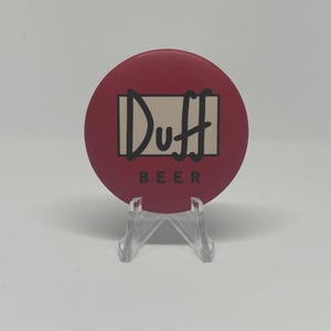 May include: A round, red button with the word "Duff" in black script inside a white rectangle. Below the rectangle, the word "BEER" is printed in black. The button is displayed on a clear plastic stand.