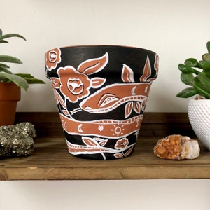 May include: A terracotta-colored flower pot with a black background and white and terracotta-colored floral and geometric designs. The pot is cylindrical and sits on a wooden shelf. The design includes flowers, leaves, and crescent moons.