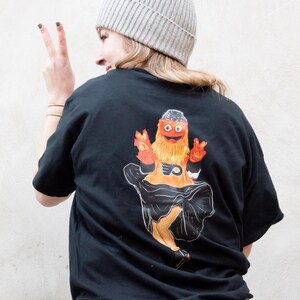 Op de afbeelding: Zwart T-shirt met een grafische print van de mascotte van de Philadelphia Flyers, Gritty, in een zwarte jurk en oranje haar. De mascotte houdt een Flyers-logo in de ene hand en een vredesteken in de andere. De persoon die het shirt draagt, draagt een grijze beanie.