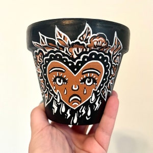 May include: Black painted ceramic flower pot with a brown heart-shaped face design. The face has a sad expression with teardrops, surrounded by white and brown floral accents. A unique decorative planter.