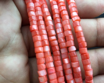 Puede incluir: Una hebra de cuentas de coral, cada cuenta es de forma cuadrada con un acabado ligeramente iridiscente. Las cuentas son de un color rojo anaranjado vibrante.