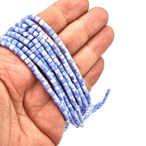 Puede incluir: Una hebra de cuentas cuadradas de color azul claro y blanco. Las cuentas están hechas de concha y tienen un acabado ligeramente iridiscente.