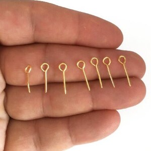 May include: A collection of small, gold-colored eye pins, each with a loop at the top and a straight pin extending downwards. The pins are arranged on a light-colored surface, showcasing their shiny, metallic finish. These are likely used for jewelry making or crafting.