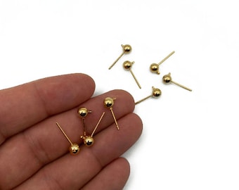 May include: A close-up of a hand holding a group of gold-colored earring findings. The findings have a small ball at the top and a straight post.