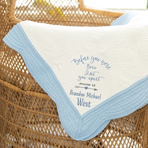 May include: A white baby blanket with a light blue border and embroidered text. The text reads "Before you were Born I set you apart Jeremiah 1:5 Brandon Michael West". The blanket is placed in a wicker basket.