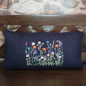Peut inclure: Un coussin en lin noir avec des fleurs sauvages brod&eacute;es dans des tons de rose, orange, bleu et vert. Le coussin est sur un banc en bois.