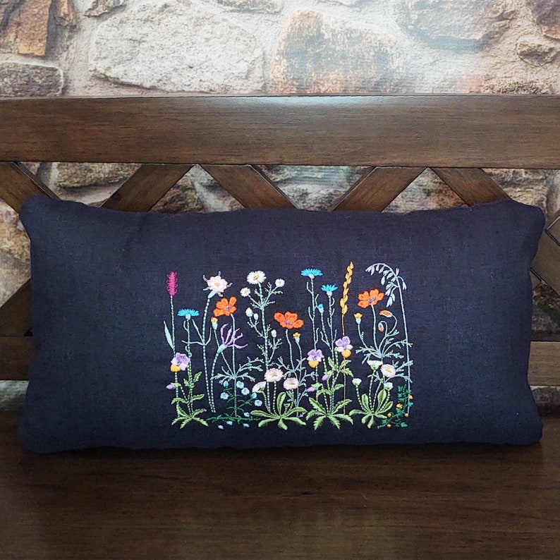 Peut inclure: Un coussin en lin noir avec des fleurs sauvages brod&eacute;es dans des tons de rose, orange, bleu et vert. Le coussin est sur un banc en bois.