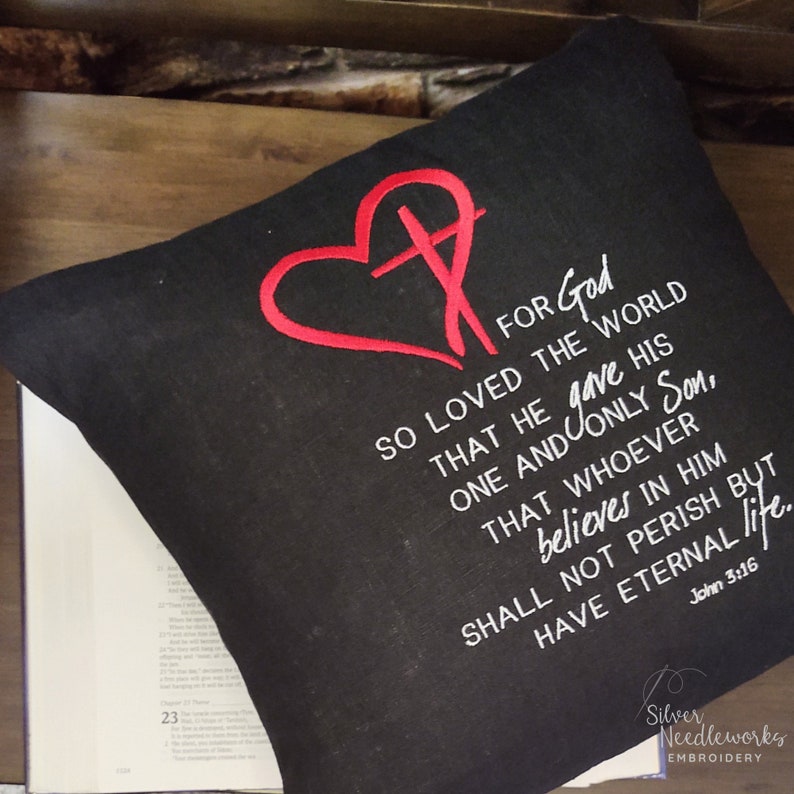 May include: Black throw pillow with white embroidery of a heart with a cross inside and a bible verse: "For God so loved the world that he gave his one and only Son, that whoever believes in him shall not perish but have eternal life. John 3:16"