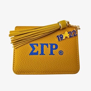 May include: Mustard yellow cardholder with blue Greek letters and a tassel. The cardholder has a textured surface and a gold-colored charm with the numbers "19 22". The tassel is also mustard yellow.