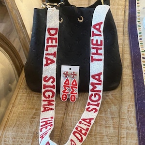 May include: Black handbag with a beaded strap featuring the text "DELTA SIGMA THETA" in red. Matching red and white earrings with the letters "OMD" are also displayed. The bag has a top handle and gold-colored hardware.