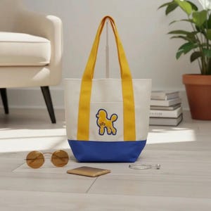 May include: A canvas tote bag with a white body, blue base, and yellow handles. A yellow poodle graphic is on the front pocket. The bag is sitting on a wooden floor with sunglasses and a wallet nearby.