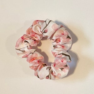 Könnte beinhalten: Ein weißer und rosa Haargummi mit Blumenmuster. Der Haargummi ist aus Stoff und hat eine gefaltete Optik.