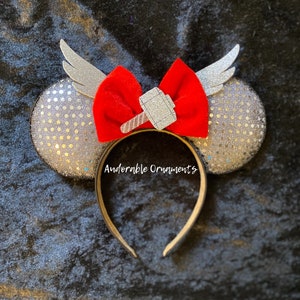 Pode incluir: Orelhas de Minnie Mouse de glitter prateado com um laço vermelho e um martelo de glitter prateado. As orelhas têm asas prateadas nas laterais.