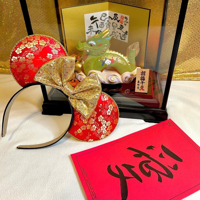 Pu&ograve; includere: Orecchie di Minnie Mouse rosse e dorate con un motivo floreale. Le orecchie sono decorate con un grande fiocco dorato. Lo sfondo include una carta rossa con il carattere cinese per "buona fortuna" scritto in inchiostro nero. La carta dice anche "Buon Anno Lunare".