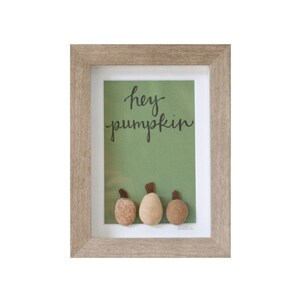 May include: A framed artwork with a green background and the text "hey pumpkin" in black script. Three small, round, brown and beige stones are arranged in a row at the bottom of the frame, resembling pumpkins.