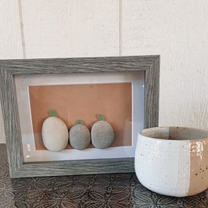 May include: A gray picture frame with a white mat and a brown background. The frame contains three smooth, round stones with green leaves on top. The stones are arranged in a row, with the largest stone on the left and the smallest stone on the right.
