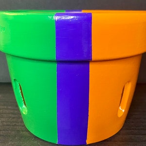 May include: A terracotta planter pot with three vertical stripes of green, purple, and orange paint. The pot has two drainage holes on the side.